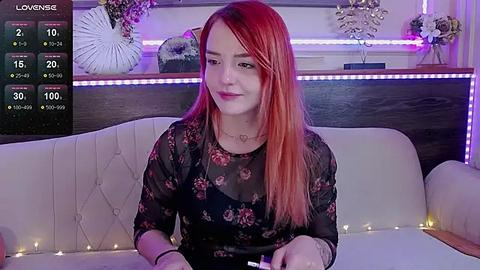 Media: Video of a fair-skinned woman with long, straight red hair, wearing a sheer black floral dress, sitting on a white couch with a wooden headrest. Background includes a dark wood wall with decorative items, purple LED strip lights, and a TV displaying live stream details.