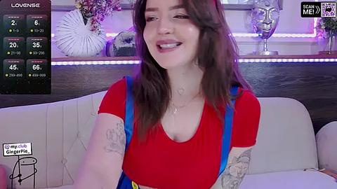 Media: Video of a smiling, tattooed woman with long brown hair, wearing a red top, seated on a white couch in a dimly lit room.