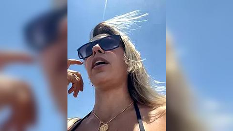 Media: A video of a tan-skinned woman with long blonde hair, wearing black sunglasses, a gold necklace, and a black bikini top, enjoying a sunny day with a clear blue sky.