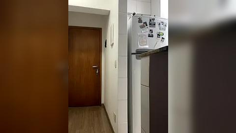 Media: Video of a modern, minimalist hallway with a wooden door on the left, white tiled walls, and a refrigerator covered in photos on the right, featuring a wooden floor.