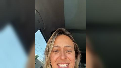 Media: Video of a smiling woman with light brown hair and a tanned complexion, seated in a car, with blurred background of trees and sky.