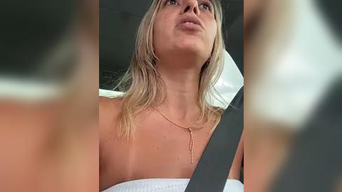 Media: Video of a blonde woman with fair skin wearing a white strapless dress and a gold necklace, looking up thoughtfully while seated in a car, with blurred faces in the foreground.