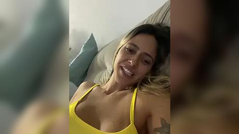 Media: A video of a smiling, light-skinned woman with medium-length blonde hair in a yellow tank top, lying on a bed with green pillows in a dimly lit room.