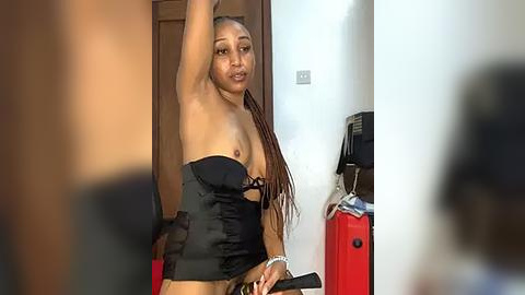 Media: Video of a Black woman with medium brown skin and long braids, wearing a black strapless corset, standing in a room with a red suitcase and white wall.