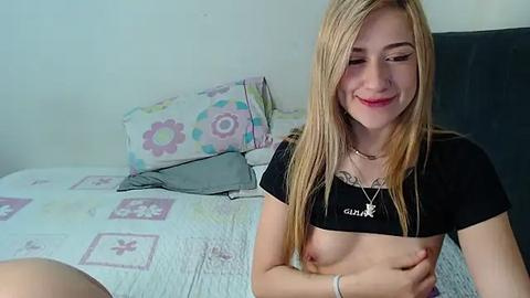 Media: Video of a smiling young woman with long blonde hair, wearing a black crop top and a necklace, revealing her small breasts, in a bed with floral-patterned sheets and pillows.