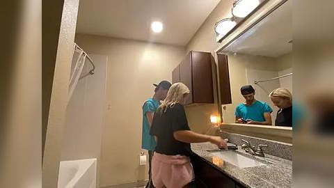 Media: Video of a bathroom with two women in teal shirts, one with blonde hair, washing hands at a granite sink. A man in a blue shirt and cap stands behind them, looking in the mirror.