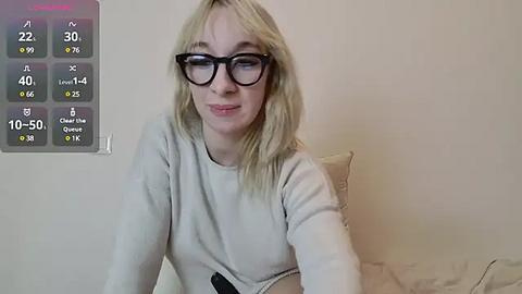 Media: Video of a smiling, fair-skinned blonde woman with glasses, wearing a light-colored sweater, sitting on a beige couch. A digital thermometer screen with temperature readings is mounted on a white wall.