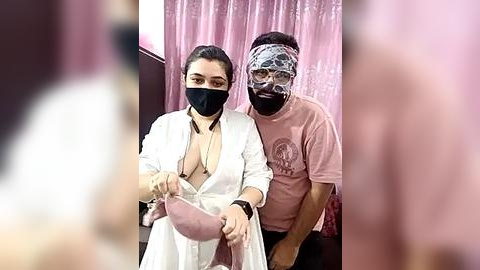 Media: Video of a woman with fair skin, dark hair, and a black face mask, wearing a white robe, holding a pink bra, standing beside a man with a black face mask, wearing a pink T-shirt, in a pink draped room.