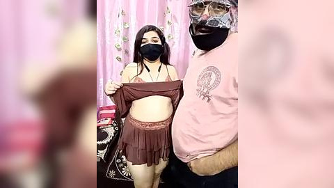 Media: Video of a masked Asian woman in brown lingerie and short skirt lifting her shirt, standing next to a masked man in a pink shirt and glasses, in a pink, floral-patterned room.