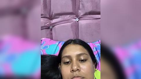 Media: Video of a young woman with medium brown skin and straight black hair, lying on a black massage table with colorful pillowcases, wearing a lavender robe, in a dimly lit room.