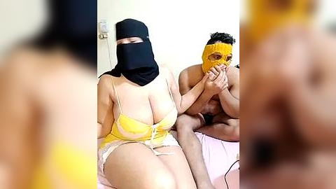 Media: Video: A light-skinned woman in a yellow bra and panties, black mask, and veil sits next to a muscular, dark-skinned man in a yellow mask, holding her hand.