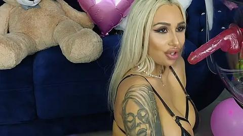 Media: Video of a blonde, tanned woman with large breasts, wearing a black harness, posing seductively on a blue couch. Background includes stuffed animals, balloons, and a dildo.