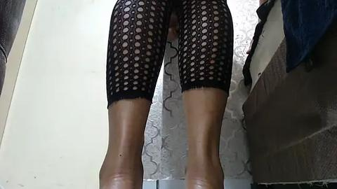 Media: Video of a person with a light brown complexion, wearing black polka dot fishnet stockings and beige high heels, standing in a bathroom with a textured white wall and a brown tile floor.