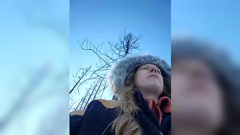 Media: Video of a blonde woman with a light complexion, wearing a furry hooded jacket, looking up at the sky with bare trees in the background against a clear blue sky.