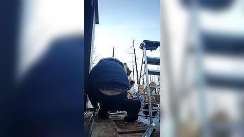 Media: Video of a person in a dark blue hoodie and jeans, squatting on a ladder, working on a boat's deck with a blue tarp covering the boat's stern. The background features a clear blue sky and bare trees.