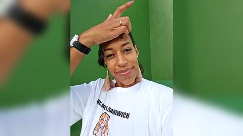 Media: Video of a woman with dark skin and short hair, wearing a white \"Hamburger University\" t-shirt, wiping sweat from her forehead, set against a green background.