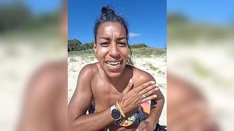 Media: Video of a joyful, tanned woman with a high ponytail, wearing a colorful bikini top, laughing and clapping on a sandy beach with green shrubbery in the background.