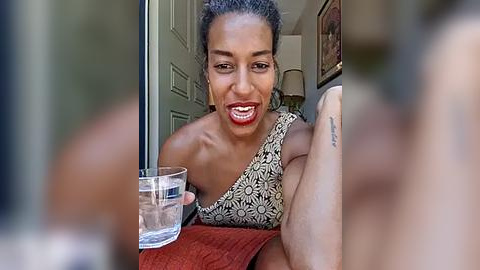 Media: Video of a smiling woman with dark skin and short hair, wearing a black and white patterned top, holding a glass of water. Background shows a hallway with closed doors and framed artwork.