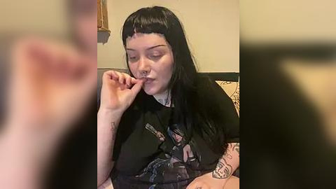 Media: Video of a pale-skinned, tattooed woman with long black hair and blunt bangs, wearing a dark t-shirt with a graphic design, biting her finger seductively indoors.