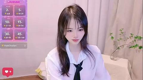 Media: A video of an Asian woman with long black hair, wearing a white blouse and black tie, seated in a living room with minimalistic decor.