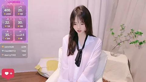 Media: A video of an East Asian woman with long black hair, wearing a white shirt and black tie, sitting on a bed with a white sheet, next to a plant.