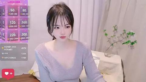 Media: A video of a young Asian woman with fair skin and dark hair in a V-neck grey sweater, sitting on a bed with white sheets and a small green plant. The image includes a virtual reality overlay with live stats and a heart icon.