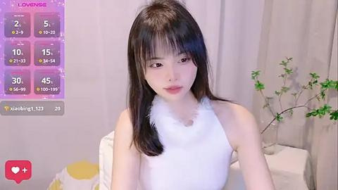 Media: Video of a young Asian woman with straight black hair, wearing a white sleeveless top with a fluffy collar, in a minimalistic bedroom setting with a green plant and white curtains.