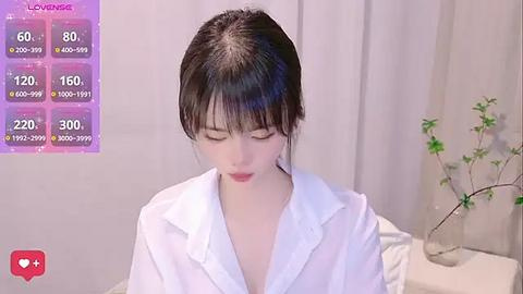 Media: A video of a young Asian woman with pale skin, dark hair, and wearing a white shirt, set in a minimalist room with a potted plant and soft lighting.