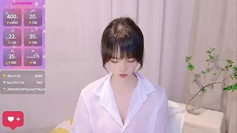 Media: Video of a young East Asian woman with fair skin and dark hair, wearing a white shirt, standing indoors near a potted plant and white curtains.