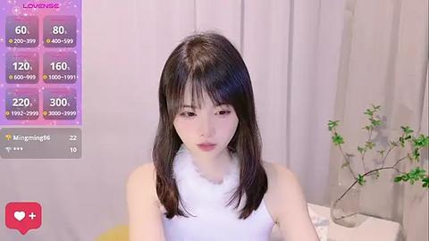 Media: A video of an East Asian woman with straight, shoulder-length black hair, wearing a white sleeveless top, indoors with a simple beige curtain background.