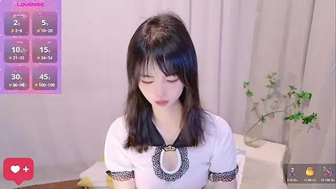 Media: Video of a young East Asian woman with straight black hair, wearing a white blouse with a black lace collar, standing in a softly lit room with a potted plant.