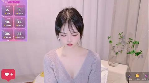 Media: A video of a young Asian woman with fair skin and dark hair, wearing a light gray sweater, sitting in a minimalist room with beige curtains and a potted plant.