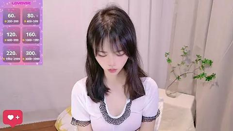 Media: A video of a young Asian woman with straight black hair, wearing a white blouse with lace trim, sitting in a room with a wooden floor, a plant, and a white curtain.