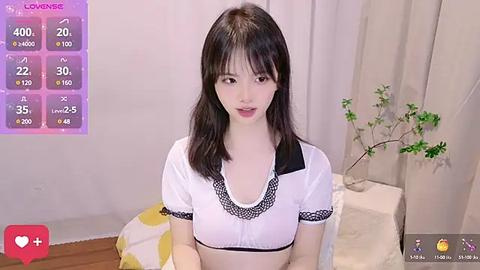 Media: A video of an East Asian woman with straight, black hair and fair skin, wearing a white crop top with black lace trim, sitting on a chair in a room with a potted plant and curtains.