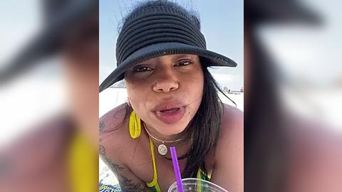 Media: Video of a young Black woman with medium-brown skin, wearing a yellow bikini, purple sunglasses, and a black bucket hat. She has tattoos on her arms and shoulders, and is smiling at the camera. Background is blurry, suggesting a beach setting.