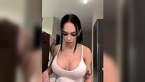 Media: Video of a young woman with pale skin, black hair, wearing a white tank top, tattoos on her arms, standing in a hallway, looking down.