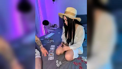 Media: A video of a woman with long black hair, wearing a straw hat and white dress, playing poker with a man in a grey shirt and shorts. The room is dimly lit with purple and blue lights.