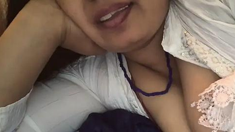 Media: Video of a partially visible, light-skinned woman with a fair complexion, wearing a white lace dress and a purple beaded necklace, lying on a bed. Her lips are slightly parted, and her arm is draped over the bed.