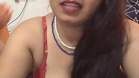 Media: Video of a smiling, light-skinned Asian woman with long, dark hair, wearing a red lace bra and two beaded necklaces. Background includes a white wall and a wire frame.