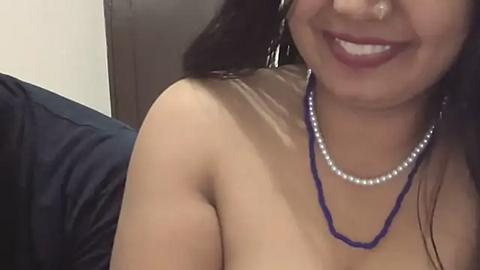 Media: Video of a smiling woman with medium skin tone, wearing a blue beaded necklace and pearl necklace, topless, dark hair, partially visible face, indoor setting.