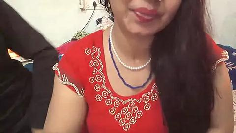 Media: Video of a South Asian woman with long dark hair, wearing a red blouse adorned with intricate white embroidery and a blue necklace, smiling. Background shows a cluttered room with a white wall and a black garment hanging.