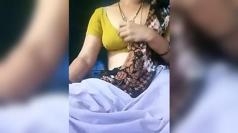 Media: Video of a woman with a medium complexion, wearing a yellow crop top, black floral skirt, and a silver necklace, sitting on a light blue blanket. The background is blurred, making the woman the focal point.