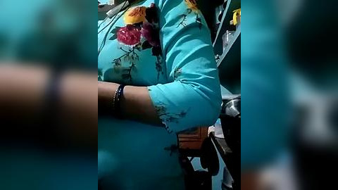 Media: Video of a person in a turquoise shirt, with a floral design, seated in a vehicle. The image is slightly blurred, focusing on the person's arm and upper torso.