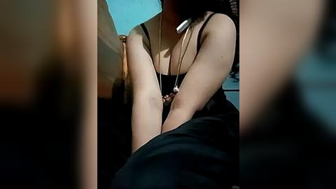 Media: Video of a light-skinned woman in a black tank top and necklace, sitting on a bed with a teal wall behind her.