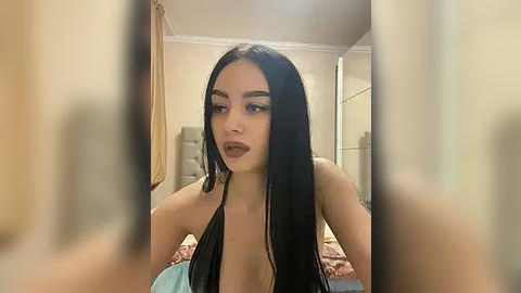 Media: Video of a young woman with long black hair, fair skin, and dark makeup, wearing a revealing black top, seated in a beige room with cream-colored walls and a bed.