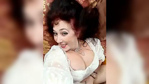 Media: A vintage video of a Caucasian woman with curly dark hair, wearing a white puffy-sleeved blouse and pearls, smiling, lying on a textured, golden-brown fabric.