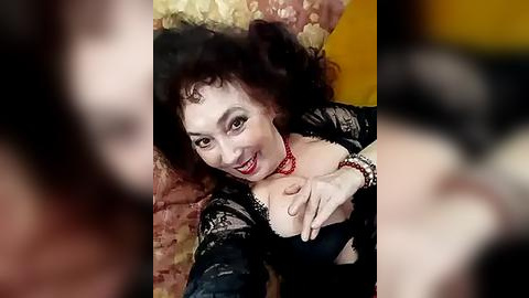 Media: A video of a woman with curly black hair, fair skin, and dark eyes, wearing a black lace bra and red choker, lying on a patterned cushion, smiling.