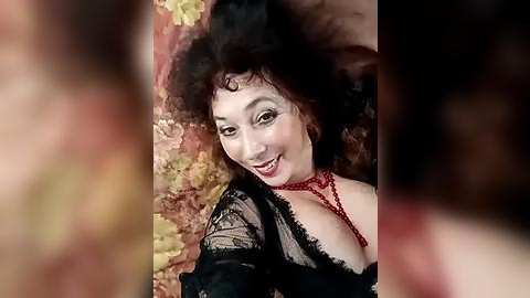 Media: A video of a fair-skinned woman with curly dark hair, wearing a black lace top, red necklace, and red lipstick, smiling against a floral-patterned background.