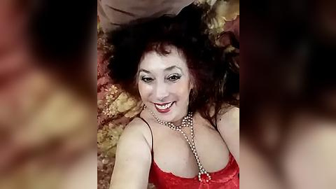 Media: A video of a smiling woman with dark hair, fair skin, and red lipstick, lying on a bed with floral pillows. She wears a red lace dress and a pearl necklace.