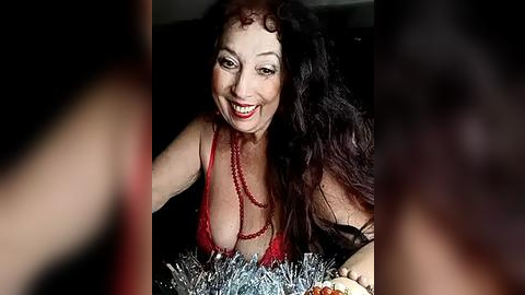 Media: Video of a smiling, middle-aged woman with long, curly black hair, wearing a red, low-cut top and silver tinsel, set against a dark background.